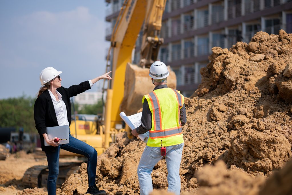 Construction managers wearing hard hats reviewing plans near heavy equipment at a noisy job